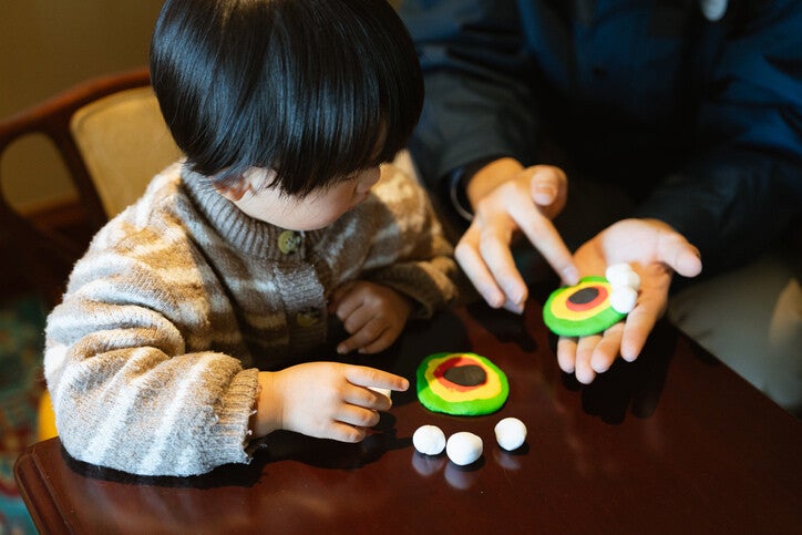 เด็กน้อยกำลังเล่นดินน้ำมันปั้นเป็นรูปทรงกลมหลากสีสันร่วมกับผู้ปกครองอย่างตั้งใจ